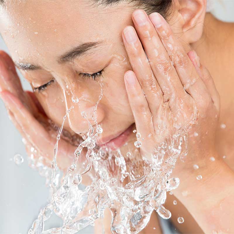 Imagem em close do rosto de uma mulher asiática. Ela possui pele branca e está com os olhos fechados, enquanto lava seu rosto. Suas mãos estão posicionadas nas laterais da face e à sua frente é possível visualizar a água que está escorrendo por ente seus dedos.