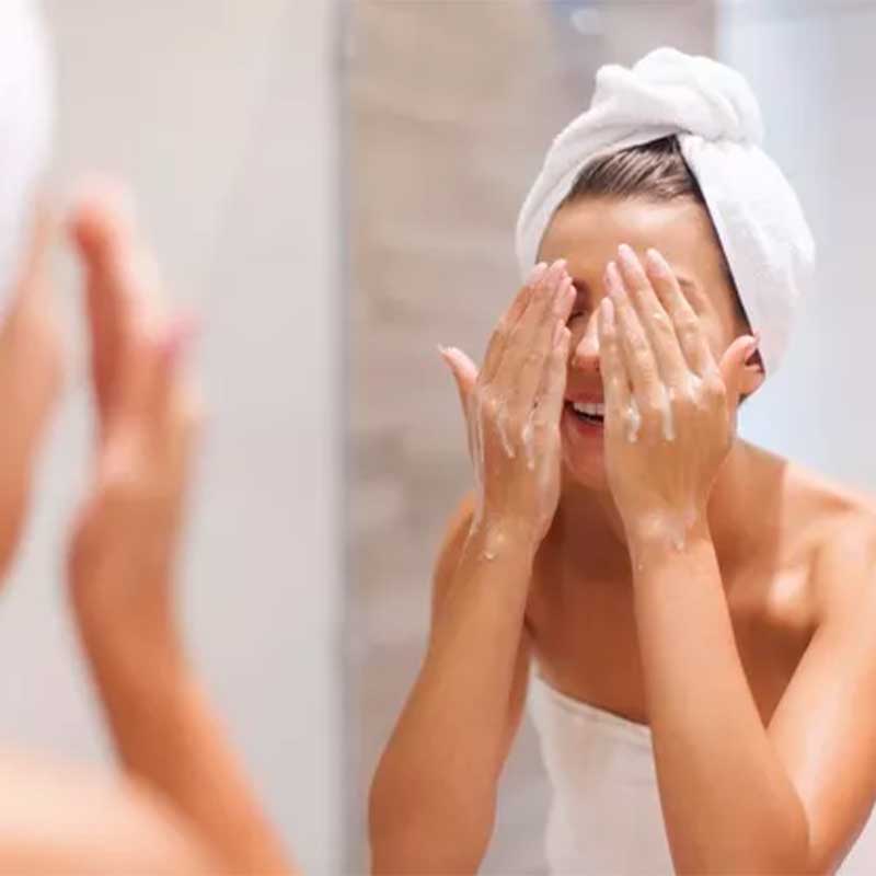 A imagem mostra uma mulher branca em frente ao espelho. Ela está enrolada em uma toalha branca e seus cabelos também, e suas mãos estão posicionadas sobre seus olhos. O fundo da imagem tem tom claro e plano embaçado.