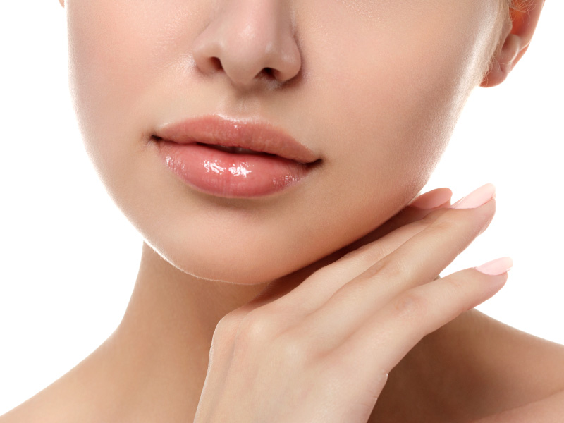 A imagem mostra, em close, o rosto de uma mulher – do nariz ao queixo. Ela está com a mão direita posicionada abaixo do queixo, exibindo lábios volumosos após a realização do preenchimento labial. O fundo da imagem é branco.