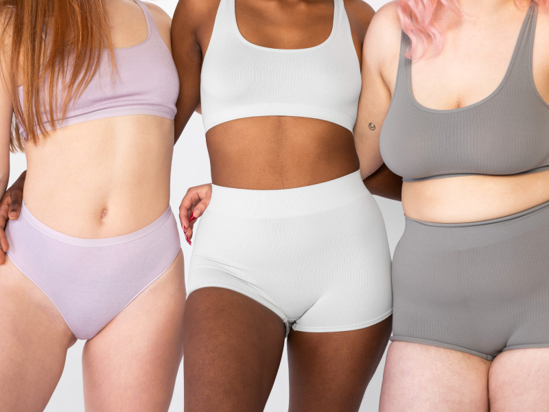 A imagem mostra três corpos femininos, posicionados lado a lado. Da esquerda para a direita, há uma mulher branca, de cabelo claro, longo e liso, vestindo calcinha e soutien rosa-claro; uma mulher negra, vestindo calcinha e soutien brancos, e uma mulher branca, de cabelo rosa, curto, na altura do ombro, vestindo calcinha e sutiã cinza.